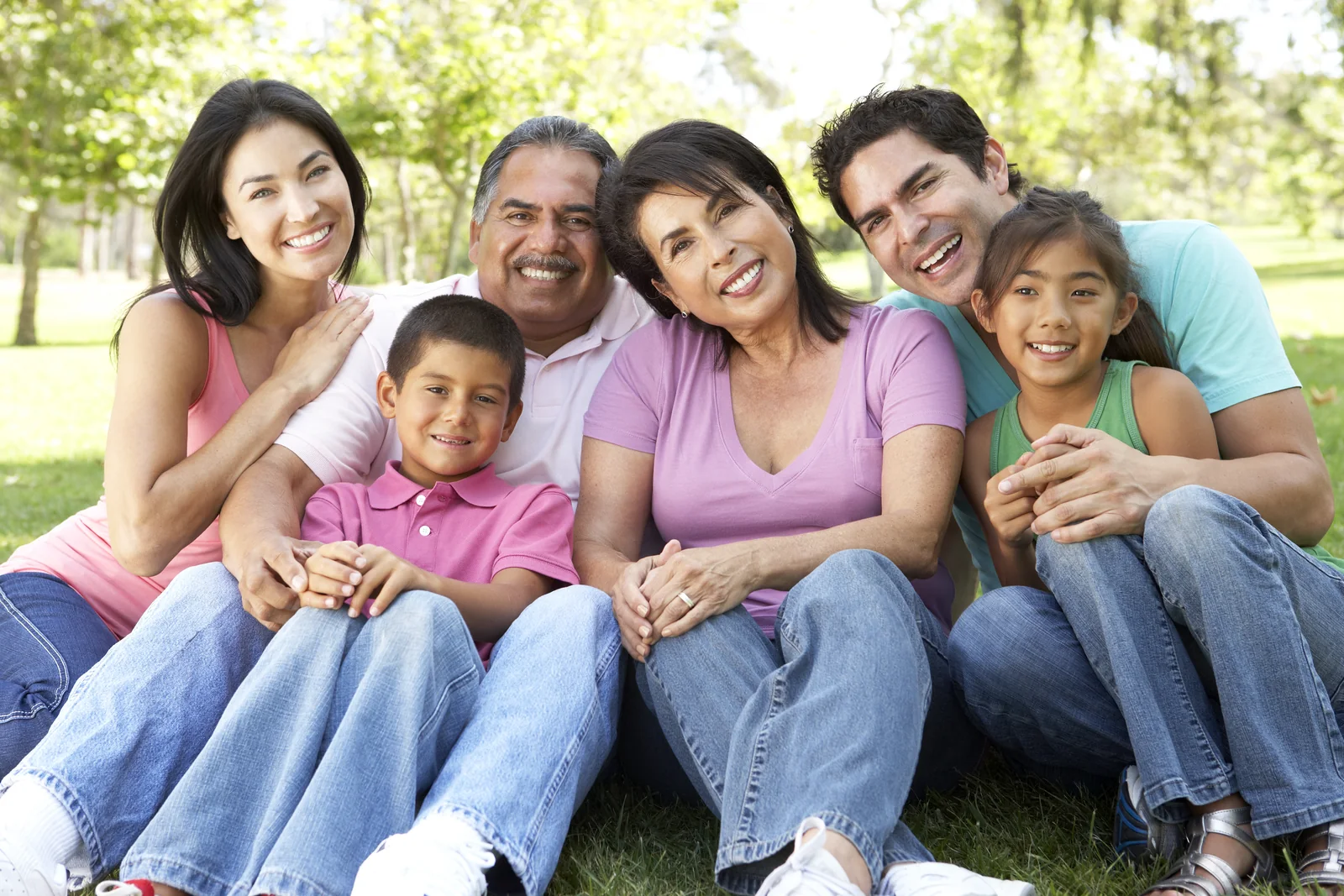 Three generations of a smiling Latino family — grandparents, parents and two children — sitting together in a park, the families Clínica Médica Guerrero cares for in Tustin